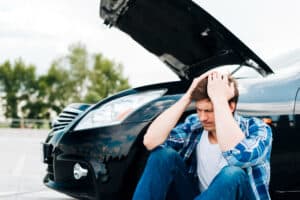 Frustrated man sitting by a broken-down car with hood open, showing vehicle trouble