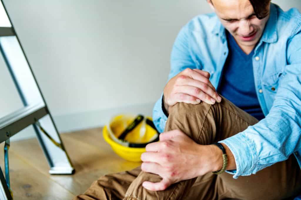 Injured worker sitting on the floor holding knee near ladder and helmet