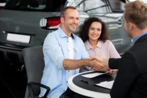 A man and woman sit at a desk, smiling, while the man shakes hands with another man across from them. A clipboard with papers and a pen are on the desk, emphasizing consumer protection and warranty law as a car is visible in the background.